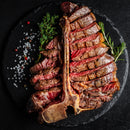 A sliced medium rare T-bone steak rests on a round black plate, garnished with rosemary and thyme, with SALTOPIA Infused Sea Salts BBQ Blend: HE MAN and peppercorns scattered beside it.