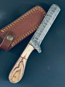 The Texan Knives Cowboy Bull Cutter Knife features a 5" Damascus steel blade with Pakkawood handle, displayed beside its brown leather sheath on a dark surface—highlighting the craftsmanship of premium handmade knives.
