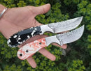 A hand displays two Texan Knives Cowboy Bull Cutter Knives with Damascus steel blades and decorative black-and-white and red-and-white handles, set against a backdrop of green leaves. Each knife comes with a leather sheath.