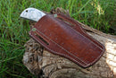 The Texan Knives Custom Handmade Cowboy Bull Cutter Knife features a 4" J2 steel blade and a white handle with blue designs. It is partially sheathed in a brown leather case with white stitching, resting on a weathered log amid green grass.