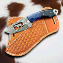 The Texan Knives Cowboy Bull Cutter Knife features a handmade Damascus steel blade with a blue-black handle, resting on a tan basketweave-stitched leather sheath against cowhide—a standout piece in the collection.