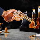 A hand holds the Craftgen 3 Gun Whiskey Decanter Set by Craftgen, made of lead-free borosilicate glass and filled with whiskey. Bullets and liquor bottles sit on a gray tabletop—an ideal centerpiece for any whiskey bar.