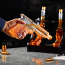 A hand holds the Craftgen 3 Gun Whiskey Decanter, a lead-free borosilicate glass bottle shaped like a handgun by Craftgen. Bullet-shaped containers and matching set pieces complete this unique whiskey bar display on a gray surface.