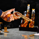 A hand holds the Craftgen 3 Gun Whiskey Decanter Set by Craftgen, featuring a revolver-shaped, lead-free glass decanter filled with whiskey; bullet-shaped glasses and bottles are arranged on the whiskey bar display in the background.