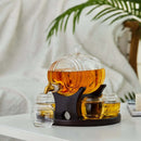 The Craftgen Barrel Whiskey Decanter Set by Craftgen features a barrel-shaped glass decanter on a dark stand, filled with amber whiskey, and is paired with four matching round glasses on a white table, with a leafy plant and draped white backdrop.