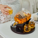 A hand holds a small glass cup of tea beside the Craftgen Barrel Whiskey Decanter Set and three cups on a round tray atop a white table, making a unique home bar centerpiece.