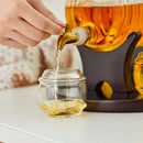 A hand pours whiskey from the golden spout of a Craftgen Barrel Whiskey Decanter Set into a small clear glass jar on a white surface.