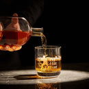 A hand pours amber whiskey from the Craftgen Baseball Decanter Set by Craftgen into a "Hit That" glass with a golfer silhouette, all placed on marble against a dark background.