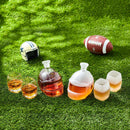 The Craftgen Football Whiskey Decanter and glasses, filled with whiskey, rest on grass beside a mini football helmet—an ideal game day gift from Craftgen for any football fan’s barware collection under dappled sunlight.