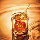 An orange ice sphere drops into a glass of amber liquid, rippling the surface—a striking way to highlight the Craftgen Golf Ball Shot Glasses - Set of 4, a must-have addition to your golf-themed barware collection from Craftgen.