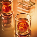 A close-up of a Craftgen Golf Ball Shot Glass filled with an amber drink and a spherical ice cube, set on a reflective surface with warm lighting—perfect for showcasing the Craftgen Golf Ball Shot Glasses - Set of 4.