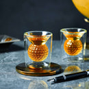 A Craftgen Golf Ball Shot Glass from the Craftgen Golf Ball Shot Glasses - Set of 4 sits on a gold coaster, its golf ball-inspired shape gleaming beside another glass and a fountain pen atop an elegant gray marble surface.