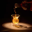 A golden-brown liquid is poured from the Craftgen Golf Liquor Decanter Set by Craftgen into a clear glass with a spherical ice ball, highlighted by warm lighting against a dark background.