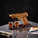 The Craftgen Gun Whiskey Decanter Set by Craftgen features a pistol-shaped decanter on a dark wood stand with two matching shot glasses, making it a unique home bar gift. A pen, cigar, and magazine rest nearby on the marble surface.