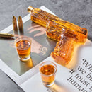 The Craftgen Gun Whiskey Decanter Set by Craftgen features a glass gun decanter filled with amber whiskey, resting on an open magazine beside two bullets, a gold pen, and shot glasses—a unique home bar gift on a gray marble surface.