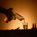 A hand pours whiskey from the Craftgen Gun Whiskey Decanter Set into a shot glass. Two bullets and a wheat stalk rest nearby, creating an ideal scene for featuring this unique Craftgen home bar gift against a warm brown gradient background.