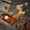 The Craftgen Golf Decanter Set by Craftgen features a club-shaped decanter filled with whiskey and a golf ball inside, surrounded by four glasses—a unique gift for golf lovers. Pen, newspaper, two white columns, and cigar box complete the stylish display.