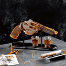 The Craftgen Gun Whiskey Decanter Set by Craftgen is shown on a marble surface, filled with whiskey and displayed on a stand alongside two shot glasses, a cigar, a bullet-shaped pen, and a magazine.