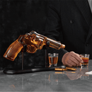 A person in a pinstriped suit holds a shot glass of whiskey next to the Craftgen Gun Whiskey Decanter Set by Craftgen, which features a gun-shaped decanter on a stand, two bullet-shaped accents, and another filled shot glass on a gray marble surface.