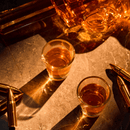 Two glasses of amber whiskey on marble, surrounded by shiny bullets, reflect warm light—a striking backdrop to highlight the Craftgen Gun Whiskey Decanter Set by Craftgen.