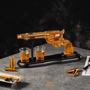 The Craftgen Gun Whiskey Decanter Set by Craftgen features a revolver-shaped decanter with amber whiskey, displayed on a wooden stand alongside two matching shot glasses and bullet-shaped accents on a reflective gray surface.