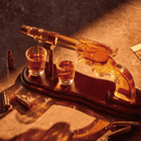 The Craftgen Gun Whiskey Decanter Set by Craftgen features a glass revolver decanter on a wooden stand, filled with amber liquid. Two whiskey glasses rest below, with gold bullets nearby—western-inspired barware for dramatic display.