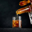 A close-up of amber whiskey from the Craftgen Rifle Whiskey Decanter Set by Craftgen being poured into a clear glass, with bubbles forming in the liquid against a dark background.