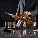 A person in a pinstripe suit stands behind the Craftgen Rifle Whiskey Decanter Set, featuring a rifle-shaped decanter on a stand with two glasses by Craftgen. A cigar and paper add flair to this distinctive barware centerpiece.