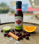 A bottle of Brushfire Farms Cranberry Pepper Sauce by Brushfire Farms, the ideal holiday condiment, rests on a barrel alongside a lime wedge, orange slice, cranberries, a cinnamon stick, and coarse salt, set against a softly blurred outdoor background.