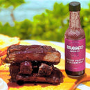 A bottle of Bravado Spice Co Ghost Pepper & Blueberry Hot Sauce from Bravado Spice Co, known for its appearance on Hot Ones Season 3, rests next to a stack of glazed BBQ ribs on an outdoor table, set against a backdrop of softly blurred green foliage.