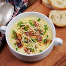 A bowl of creamy soup topped with shredded cheese, bacon bits, green onions, and a sprinkle of SALTOPIA Infused Sea Salts Chive Salt: DIVE INTO CHIVES sits on a wooden board beside bread slices and a metal spoon.