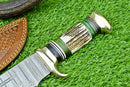 A close-up of the Texan Knives Deluxe Dundee Knife, featuring a 16" handmade Damascus steel blade, brass guard, and stag horn handle, resting on artificial grass beside its custom leather sheath with detailed stitching.