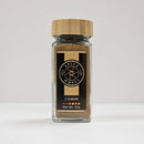 A glass jar with a wooden lid labeled "Spice Wheel Cumin Powder" by Spice Wheel, Net Wt. 57g, rests on a white surface against a plain white background, highlighting this earthy spice celebrated for its distinctive nutty flavor.