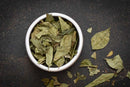 A white bowl filled with World Spice Curry Leaves sits on a dark, textured surface, with some loose leaves scattered around the bowl, showcasing this essential ingredient from World Spice, renowned for its aromatic flavor in South Indian cooking.