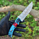 A gloved hand holds the Texan Knives Custom Handmade Damascus Steel Hunting Knife – 10-Inch Blade with a blue, red, and black resin handle, shown outdoors with a blurred green and brown background. Leather sheath included.