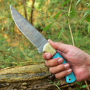 A hand holds the Texan Knives Custom Handmade Damascus Steel Hunting Knife with a 10-inch blade and resin handle, outdoors near a fallen tree and green foliage. Leather sheath included.