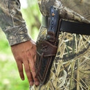 A person in camouflage stands outdoors with the Texan Knives Custom Handmade 9" Damascus Steel Hunting Knife, featuring a G10 handle and leather sheath, attached to their belt. Their hand rests beside the sheath.