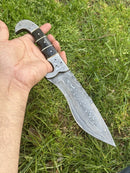 A hand holds the Texan Knives Custom Handmade 12" Damascus Steel Hunting Knife, featuring a buffalo horn handle and Damascus steel bolster, with detailed engravings on the blade, set against green grass. Includes leather sheath.