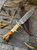 A Texan Knives Custom Handmade Damascus Steel Bowie Knife with a 13-inch blade and bone/brass guard handle rests beside its leather sheath on a stone surface, surrounded by antler pieces.