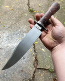 A hand holds the Texan Knives Custom Handmade 13" J2 Steel Bowie Knife with a steel guard, rosewood handle, and leather sheath, pointing downward over rough concrete with scattered leaves.