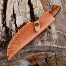 The Texan Knives Custom Handmade 5-Inch Damascus Skinning Knife with a rosewood handle is partially sheathed in a brown leather case with decorative stitching, resting on rough, textured tree bark.
