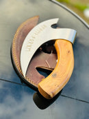 The Texan Knives Vikings Pizza Cutter – Custom Handmade 8-Inch J2 Steel Blade with a wooden handle lies on a brown leather sheath atop a glossy black surface, with runic engravings visible and an outdoor scene blurred in the background.
