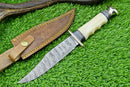 The Texan Knives Handmade Western Boulder Colorado Knife features an 11" Damascus steel blade, a camel bone handle, and a leather sheath with yellow stitching and a snap button, shown on green grass.