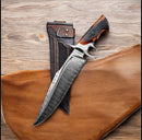 The Texan Knives Texas Knives Custom Bowie Knife, a 14" handmade Damascus steel blade with a stag horn handle, rests beside its decorative leather sheath on wood—showcasing unique markings and intricate design details. Essential for fall hunting.