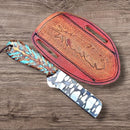 The Texan Knives Bull Cutter - Custom Handmade 8-Inch J2 Steel Knife with a turquoise-patterned handle rests beside a brown feather-embossed leather sheath, both displayed on a wooden surface.