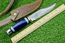 The Texan Knives Western Boulder Colorado Knife features an 11" handmade Damascus steel blade, epoxy resin handle, and leather sheath, shown beside a wooden ruler on green grass.