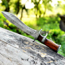 A Texan Knives Custom Handmade 13" Damascus Steel Civil War Bowie Knife with a patterned blade, rosewood handle, and leather sheath rests on a weathered log outdoors, green foliage softly blurred in the background.