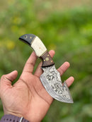 A hand holds the Texan Knives Custom Handmade Damascus Steel Guthook Skinning Knife, 7.5", featuring a rosewood, camel bone & black sheet handle, with fine craftsmanship seen against a blurred green outdoor background.