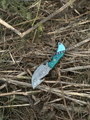 The Elite Custom Handmade Damascus Steel Guthook Skinning Knife by Texan Knives features a 7.5” premium blade and resin handle, displayed outdoors on dry sticks and grass, and comes with a leather sheath.