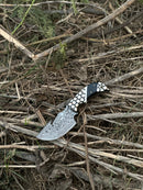 The Texan Knives Exquisite Custom Handmade Damascus Steel Guthook Skinning Knife with a unique resin handle and 7.5” blade rests outdoors on dry grass and sticks, accompanied by its leather sheath.